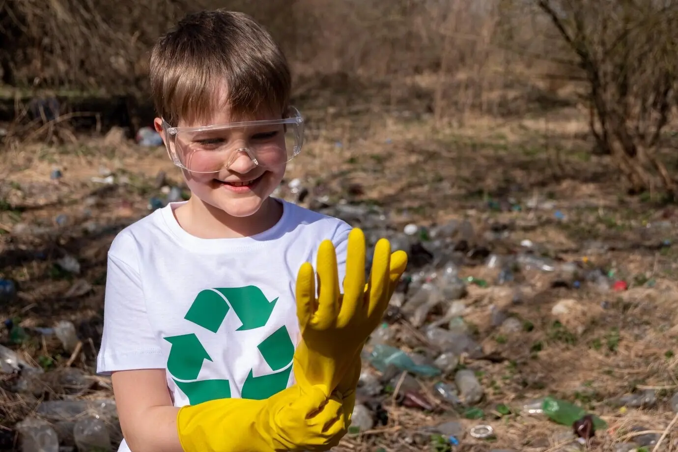 Ein junger weißer kaukasischer Junge mit einem Recycling-Symbol auf seinem T-Shirt und einer Brille, der gelbe Handschuhe anzieht.